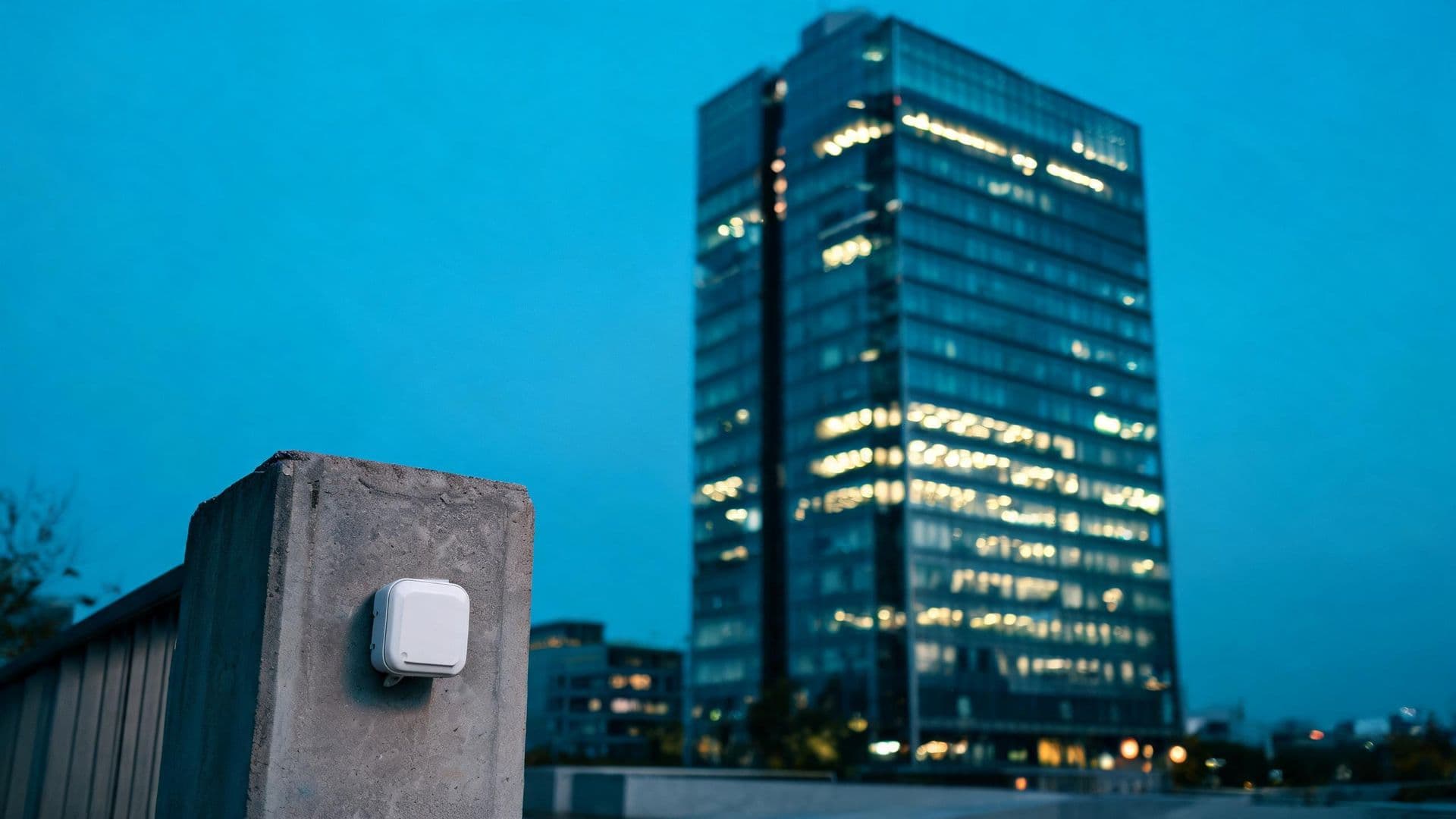 Illustrative photo: a wireless building sensor in front of a glass office tower at dusk