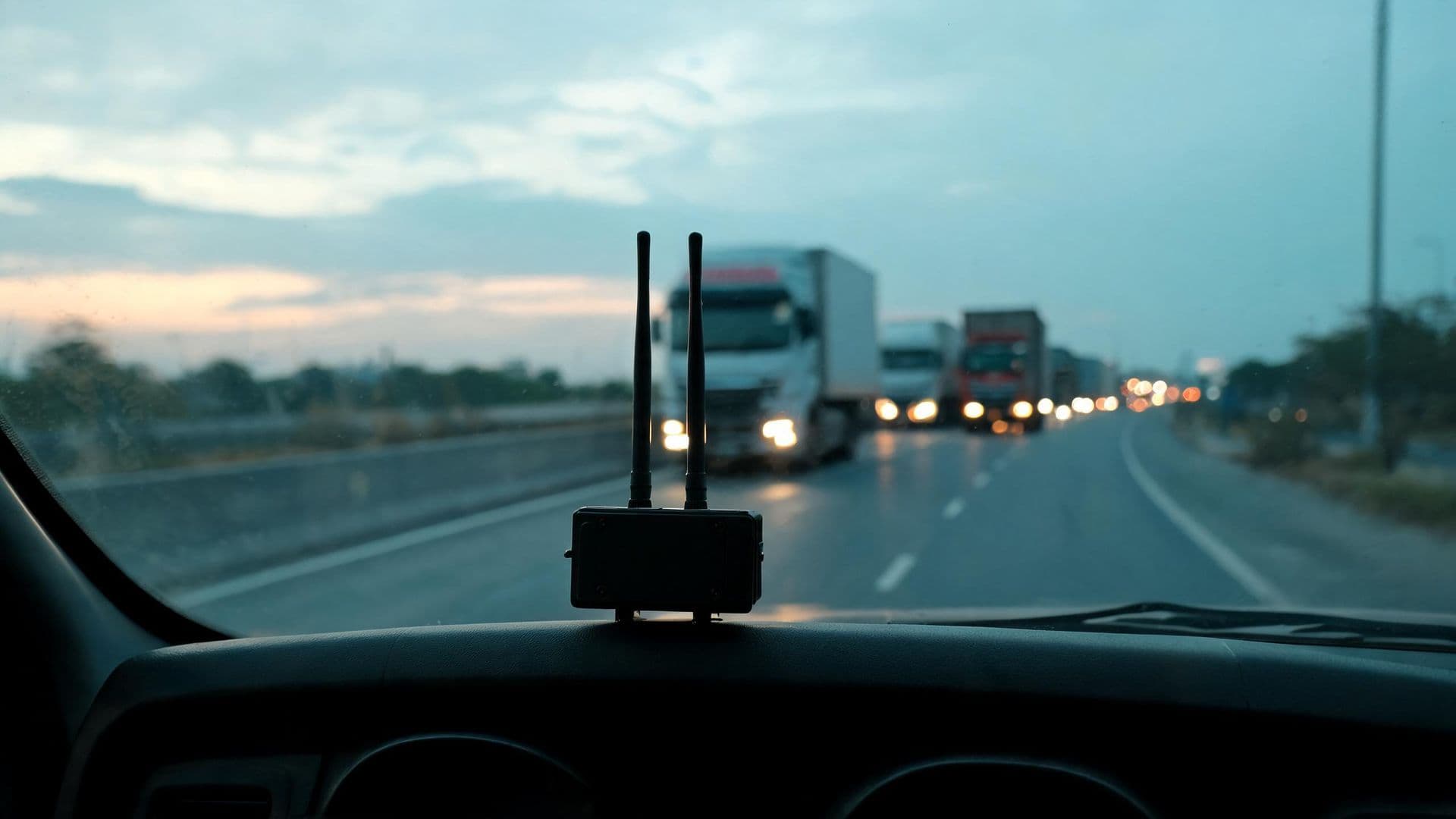Illustrative photo: a telematics unit on a truck dashboard with cargo trucks on a highway at dusk